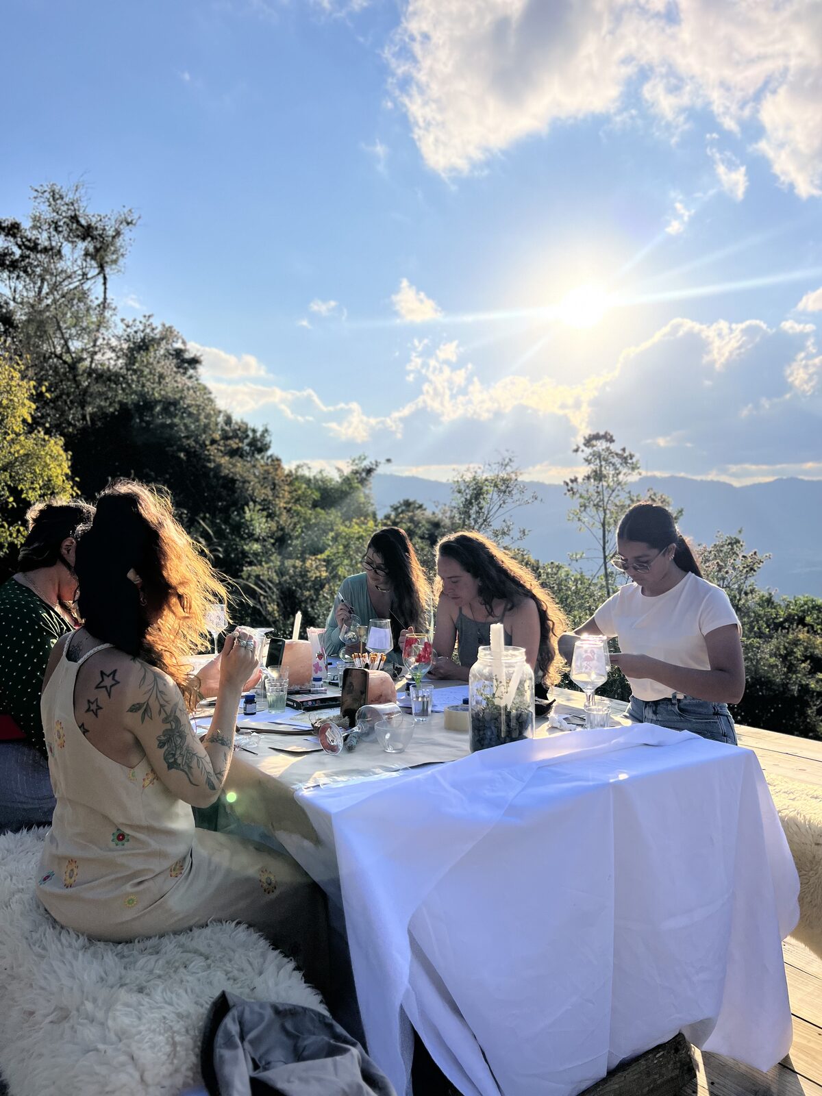 Mulheres reunidas pintando taças de vidro em uma mesa ao pôr do sol